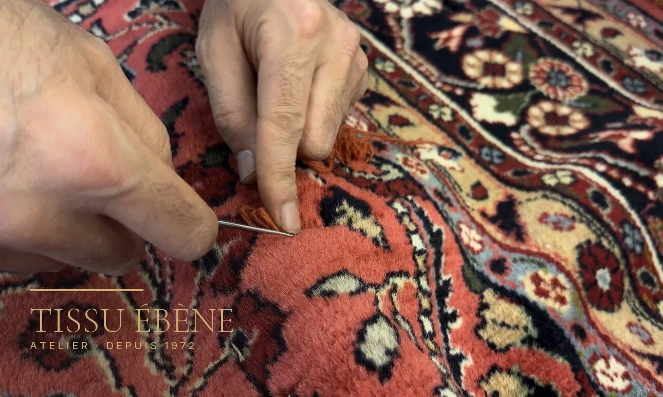 Mains tenant une aiguille et un brin de laine orangée sur un tapis noué rouge à fleurs