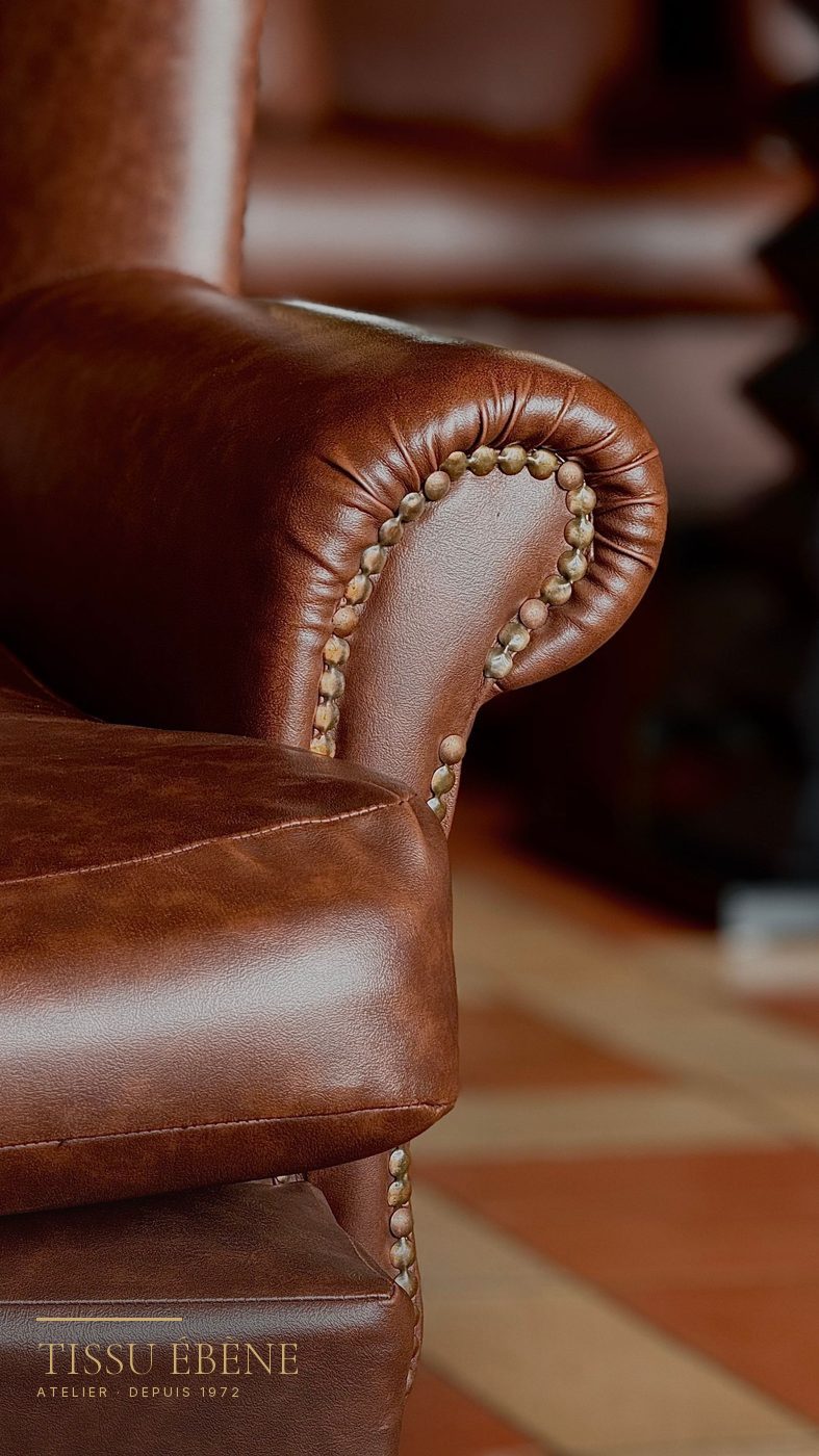Gros plan sur l&rsquo;accoudoir d&rsquo;un fauteuil en cuir brun patin&eacute;, clous de tapissier apparents