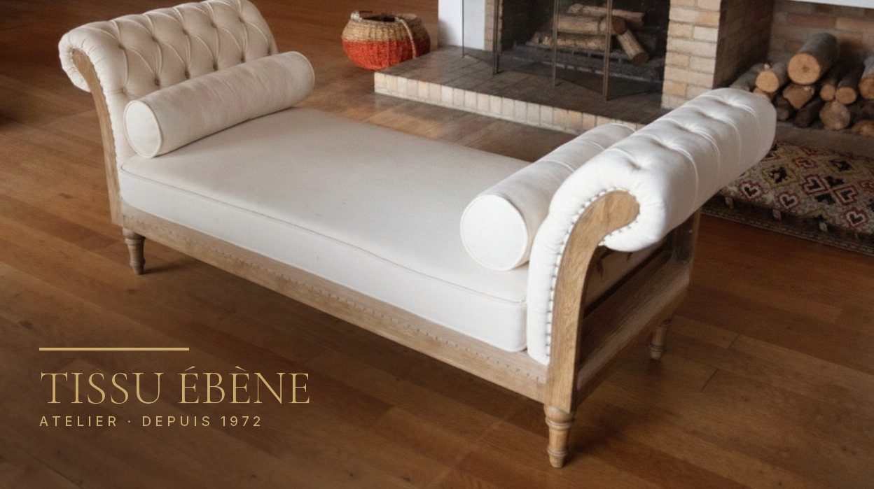 Banquette capitonn&eacute;e en tissu ivoire avec deux traversins, pi&egrave;tement en bois clair, devant une chemin&eacute;e