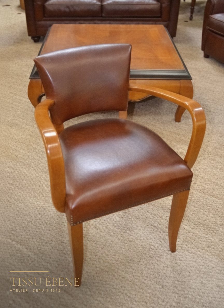 Fauteuil bridge en cuir brun et bois clair, assise clout&eacute;e, pos&eacute; sur une moquette beige