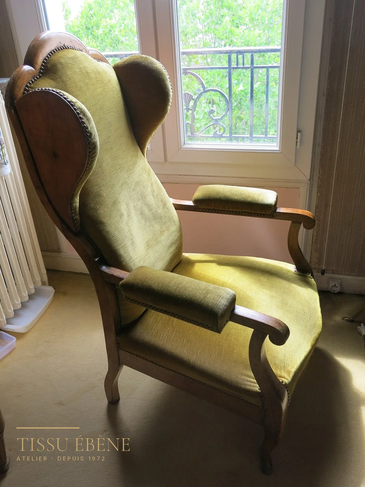 Fauteuil à oreilles ancien garni d'un velours vert olive, bois clair et clous de tapissier, devant une fenêtre