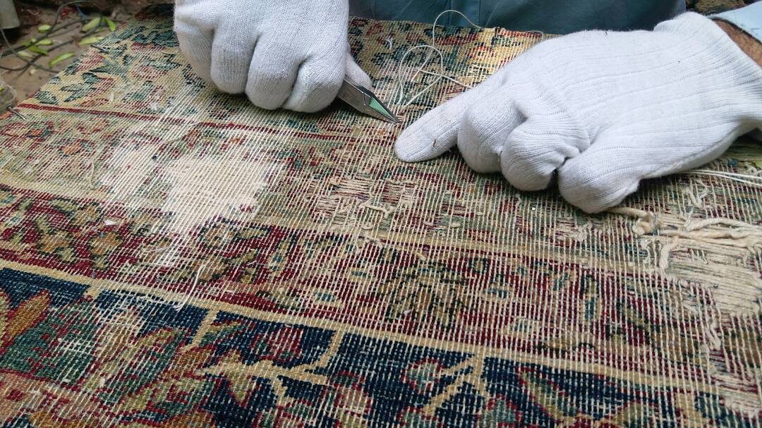 Mains gantées reprenant les fils de trame d’un tapis usé jusqu’à sa chaîne